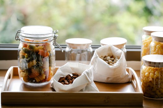 Bocaux en verre et sachets en tissu contenant des pâtes et des noix sur un plateau en bois près d'une fenêtre.