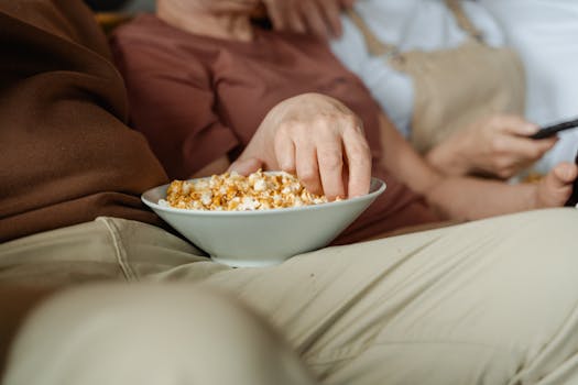 Main plongeant dans un bol de popcorn, assis sur un canapé avec d'autres personnes, ambiance de soirée cinéma.