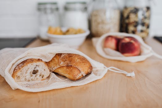 Sac en tissu contenant des pains sur une planche en bois, avec des bocaux et des fruits en arrière-plan.